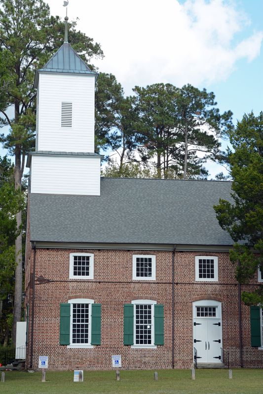 The white belfry above the grey roof and brick walls, with pines behind